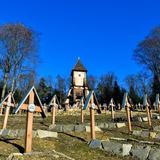 Cmentarz z kamiennymi obmurówkami i krzyżami, w oddali drewniana kaplica.