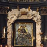 Obraz Matki Bożej Kalwaryjskiej z Dzieciątkiem, w ciemnej ramie, na jasnym tle, w ciemnych sukienkach ze złoceniami i w złotych koronach. Po bokach jasne obramowanie z aniołkami u góry podtrzymującymi koronę i napisem 