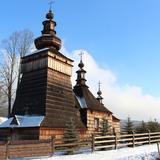 Drewniana cerkiew z dużą wieżą w zimie. Kościół ogrodzony drewnianym płotem, przy którym rosną młode drzewka choinkowe. Dach przyprószony śniegiem.