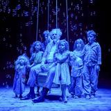 Starszy mężczyzna siedzący na podwieszonym na linach siedzisku otoczony grupką dzieci na scenie teatru w niebieskie poświacie