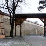 Drewniana duża brama wejściowa do klasztoru Sióstr Klarysek. Przed nią kapliczka. Za nimi wysoki mur z kamieni i wzdłuż niego droga. W bramie pachołki.
