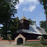 Kościół drewniany z wieżą widziany zza kamiennego ogrodzenia z bramą.