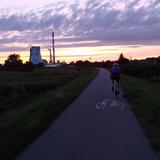 Zmrok, zachód słońca. Po wiślanej Trasie Rowerowej jedzie rowerzysta, po bokach ciemne krzewy. W oddali widać wysokie kominy.