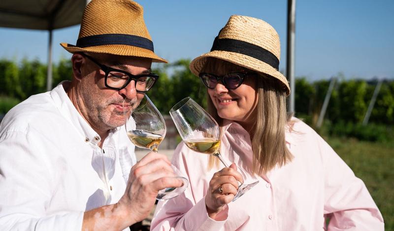 Kobieta i mężczyzna w jasnych koszulach i słomkowych kapeluszach rozkoszują się winem, w tle winnica.