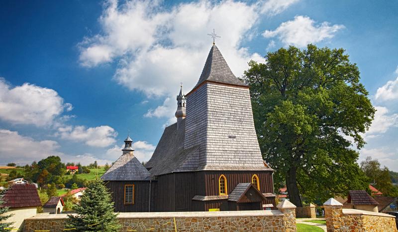 Drewniany kościół z dachem oraz wieżą pokrytymi gontem. Kościół otoczony jest kamiennym ogrodzeniem