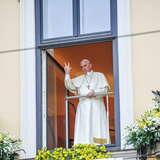 Prostokątne okno, pod nim ozdobny gzyms z donicami i kwiatami w barwach papieskich (biało-żółtych) i stojący w otwartym papieskim oknie papież Franciszek I, w białej albie, z białą piuską na głowie i błogosławiący prawą ręką.