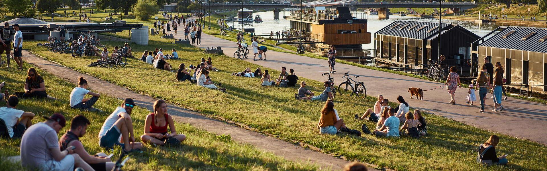 View of groups of people sitting on the grass along the Vistula Boulevards in Kraków. On the right by the shore, moored ships. In the depths, the river and the long bridge. Further on the left, trees and on the right across the river, the Wawel Royal Castle with its two round towers. Clear skies.