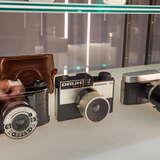 Trzy stare aparaty fotograficzne na szklanej półce w gablocie w Muzeum Fotografii w Krakowie. Za nimi widać także dwa skórzane etui.