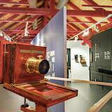 Czerwony, drewniany aparat na pierwszym planie. W dalszej części widać salę w Muzeum Fotografii w Krakowie z drewnianymi, czerwonymi elementami na suficie, a środek sali jest przedzielony ścianką.